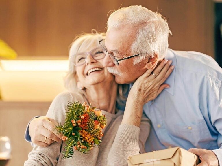 Er gratuliert ihr zu einem Jubiläum mit kleinem Blumenstrauß, Geschenk und einem Kuss.