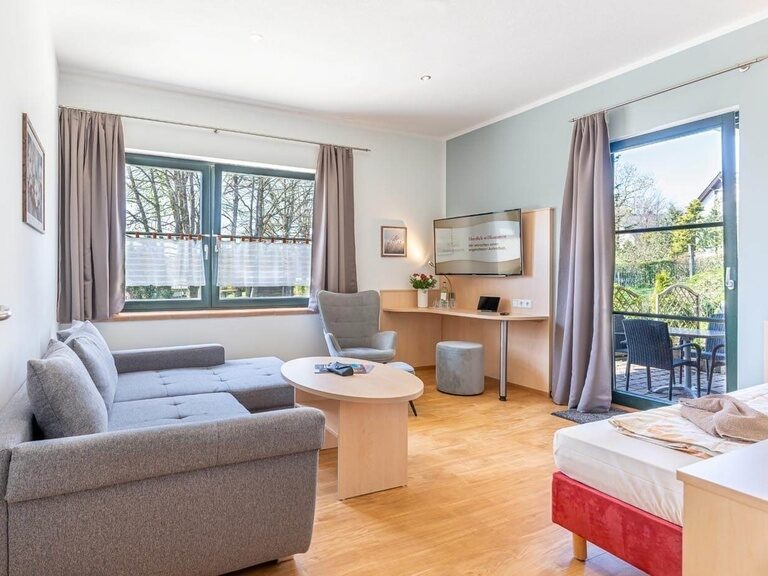 Blick in das geräumige Wohnzimmer mit Sofa, Sessel, Schreibtisch und Zusatzbett. Fenster und Terrassentür machen den Raum schön hell.