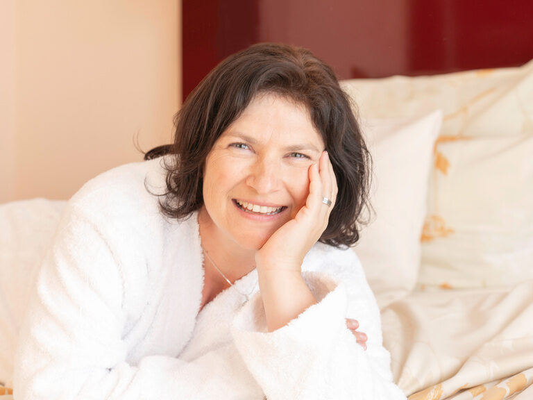 Eine Frau im Bademantel liegt auf dem Hotelbett und strahlt entspannt in die Kamera.
